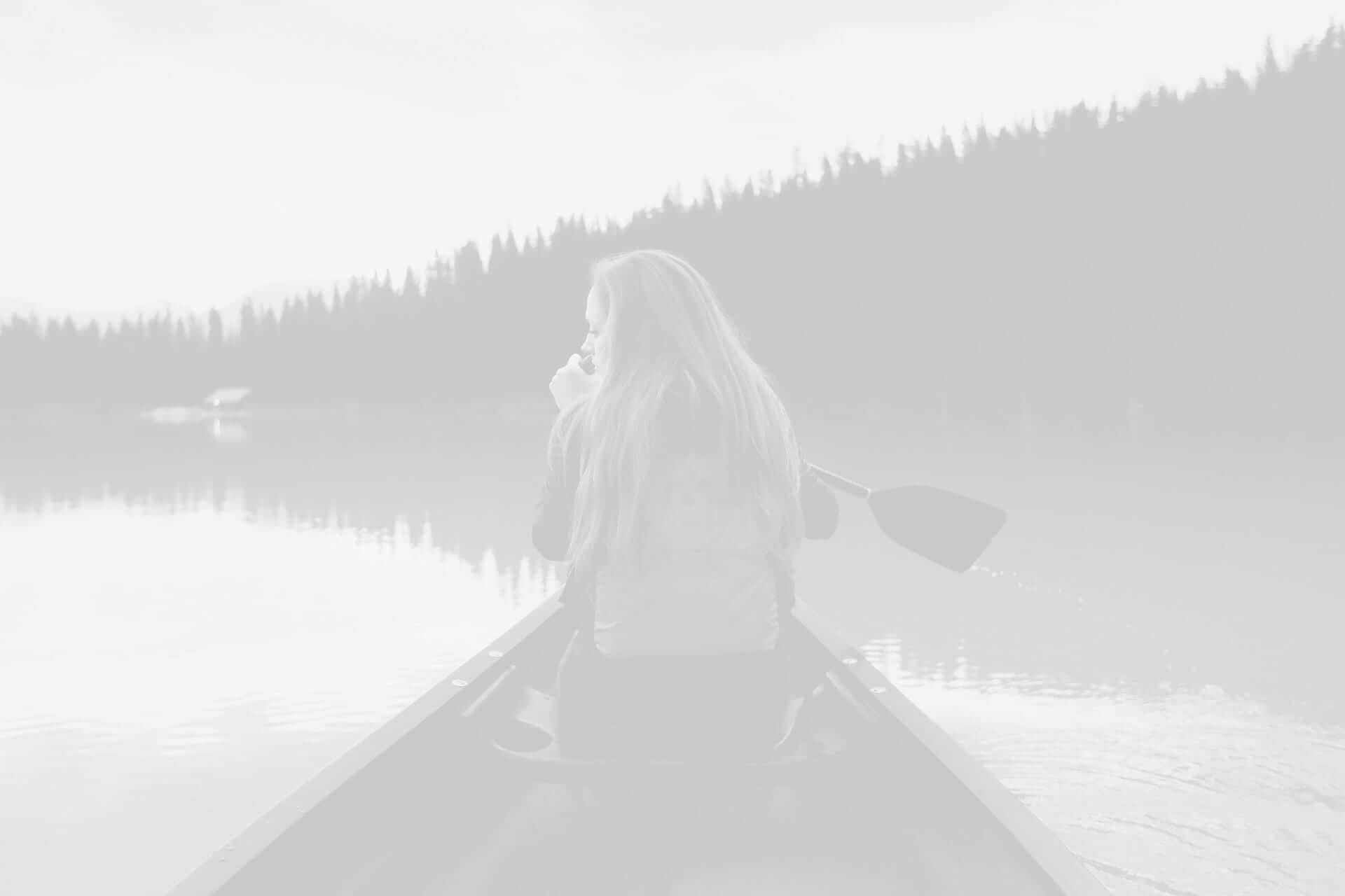 Woman sitting in a canoe on a calm lake, holding a paddle, with a backdrop of a forested hill under a cloudy sky.