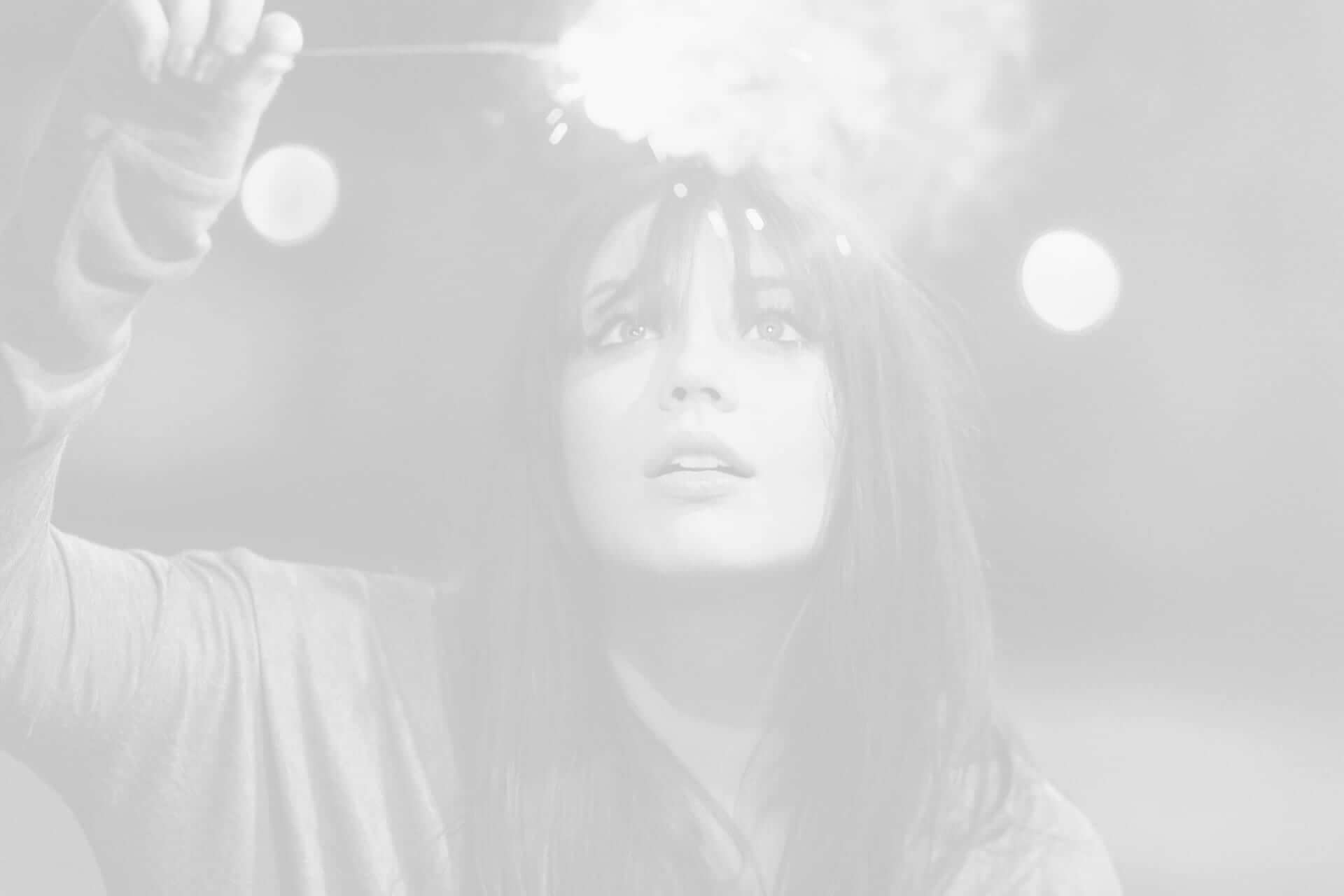 A woman holding a lit sparkler, looking upwards with a focused expression, surrounded by soft bokeh lights in a monochrome setting.