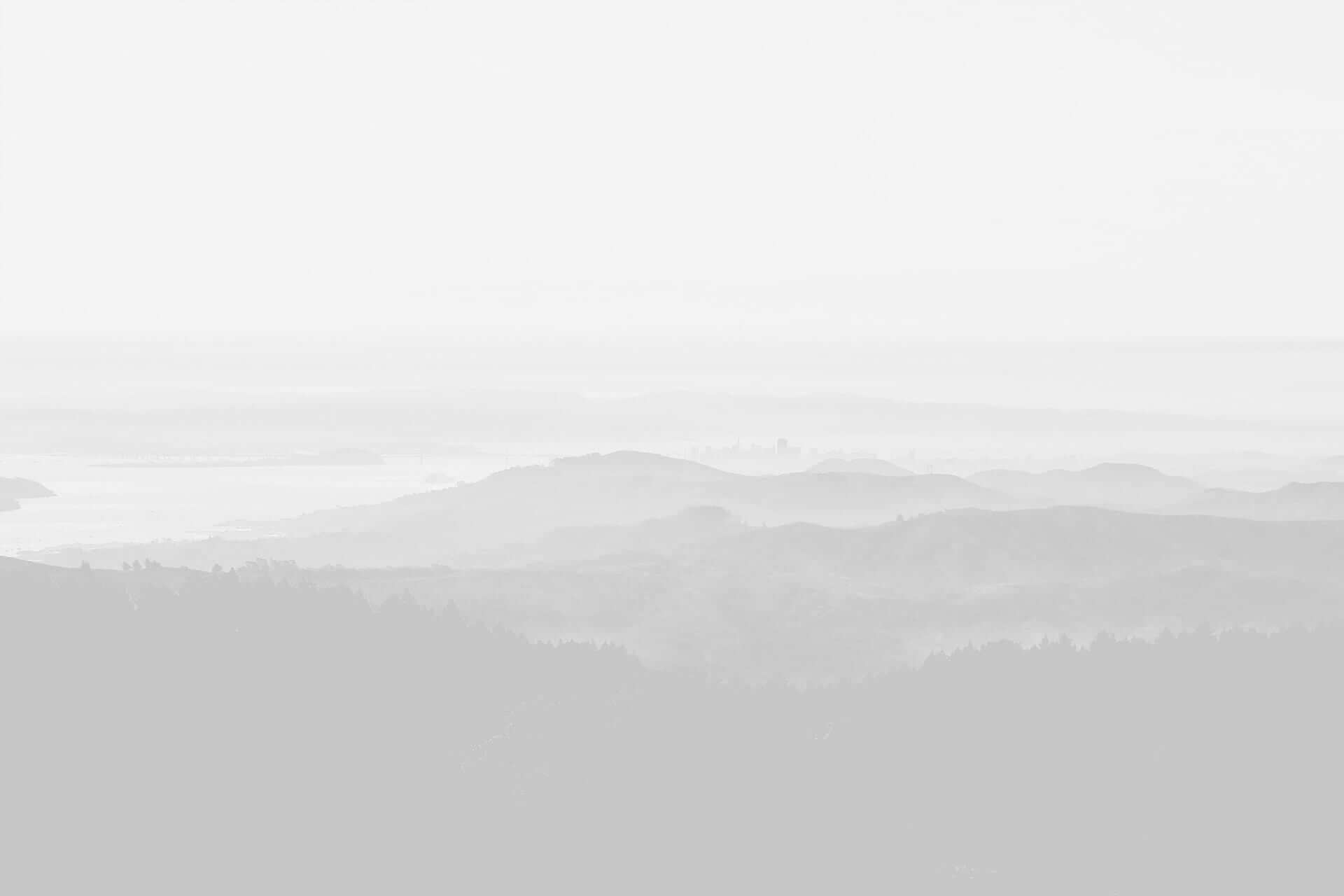 A monochrome landscape of layered mountain ridges under a hazy sky, with a hint of a water body in the distance.
