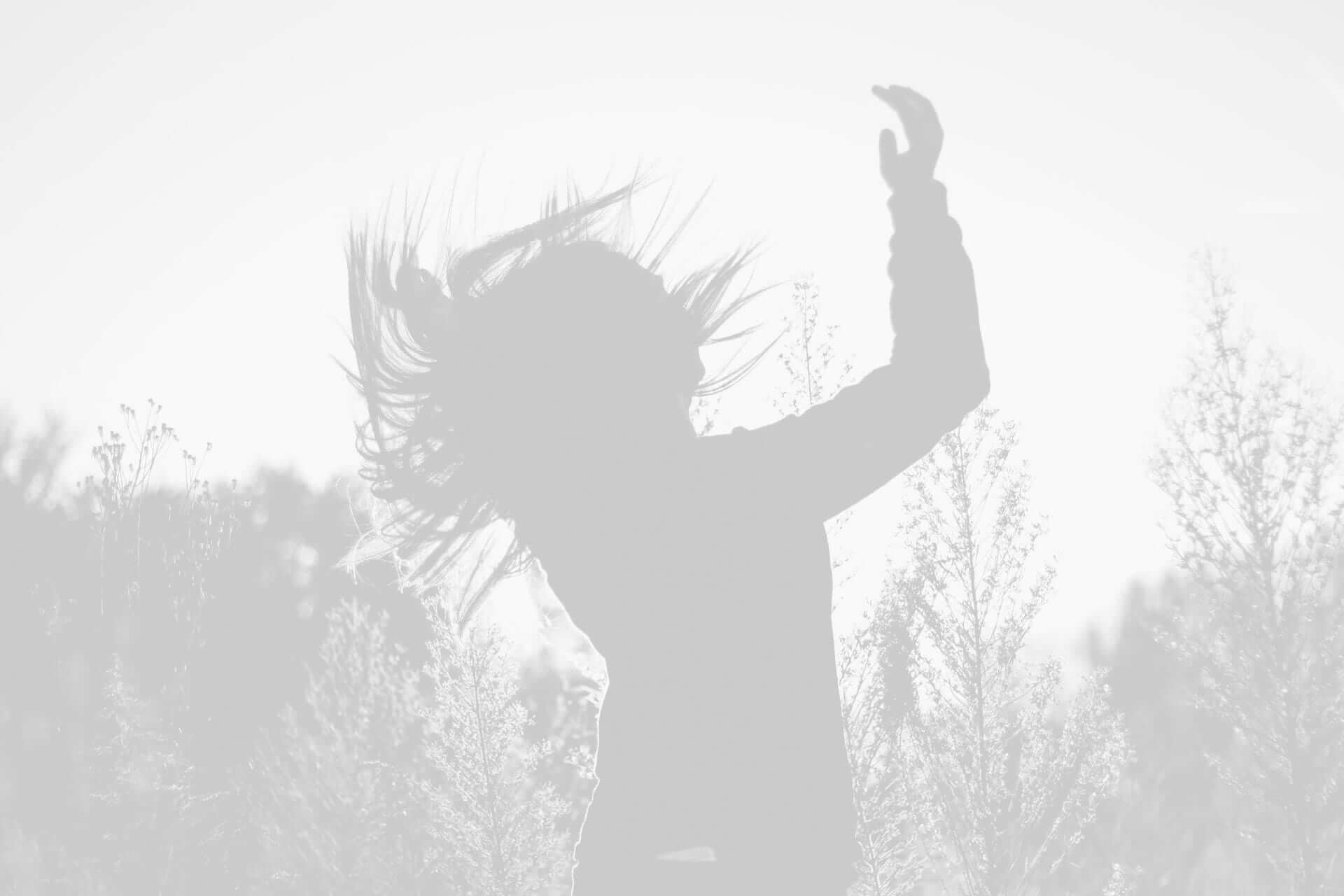 Silhouette of a woman dancing in a field, her hair blowing in the wind, backlit by a bright, diffuse light.