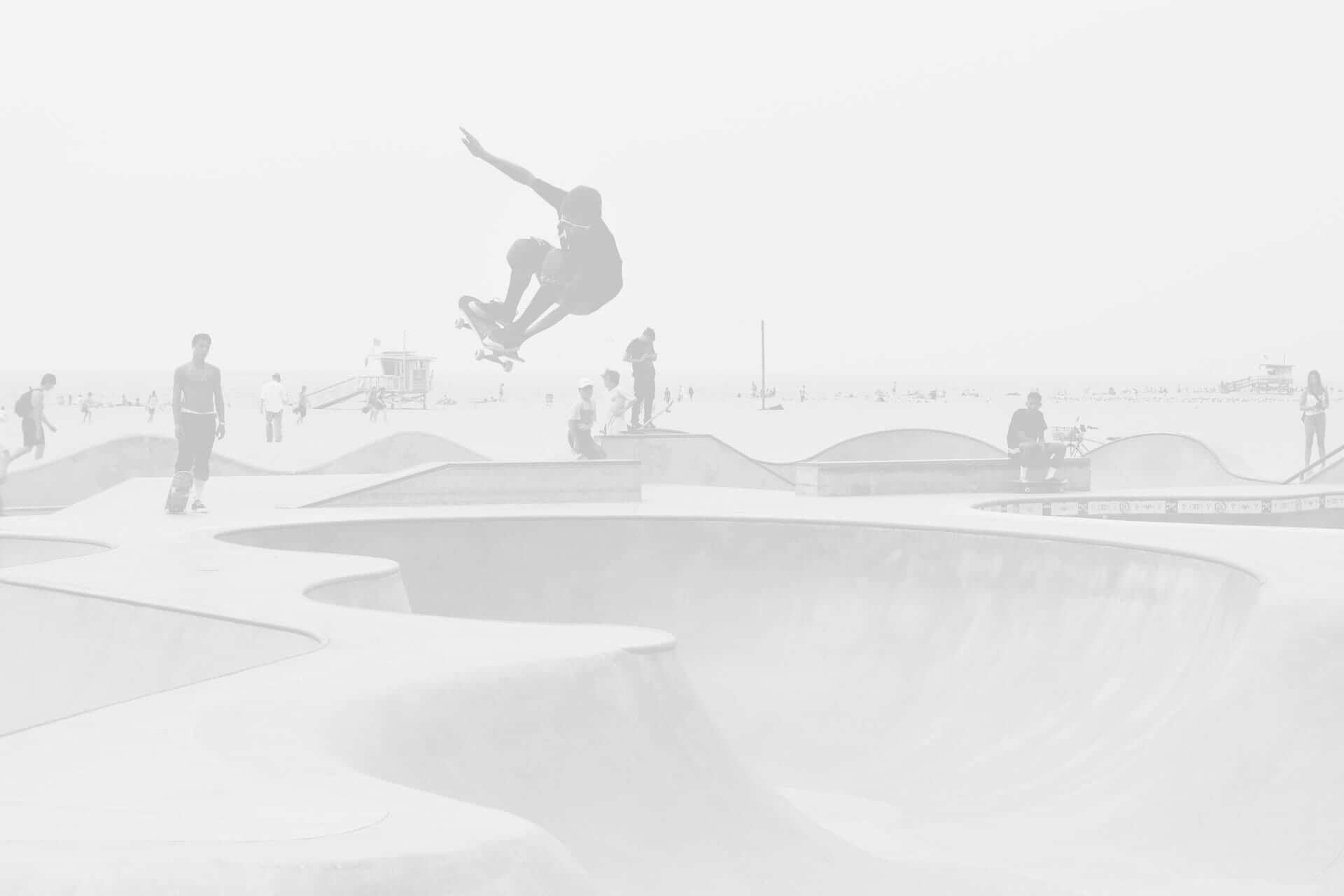 A skateboarder performs a jump in a skatepark with other skaters in the background, all in a hazy, overexposed setting.