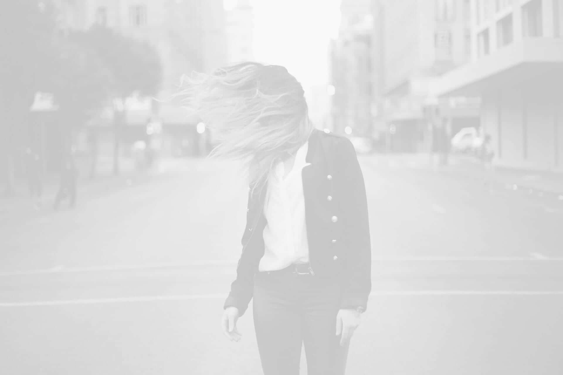 A monochrome image of a woman with her hair obscuring her face, standing in the middle of a city street.