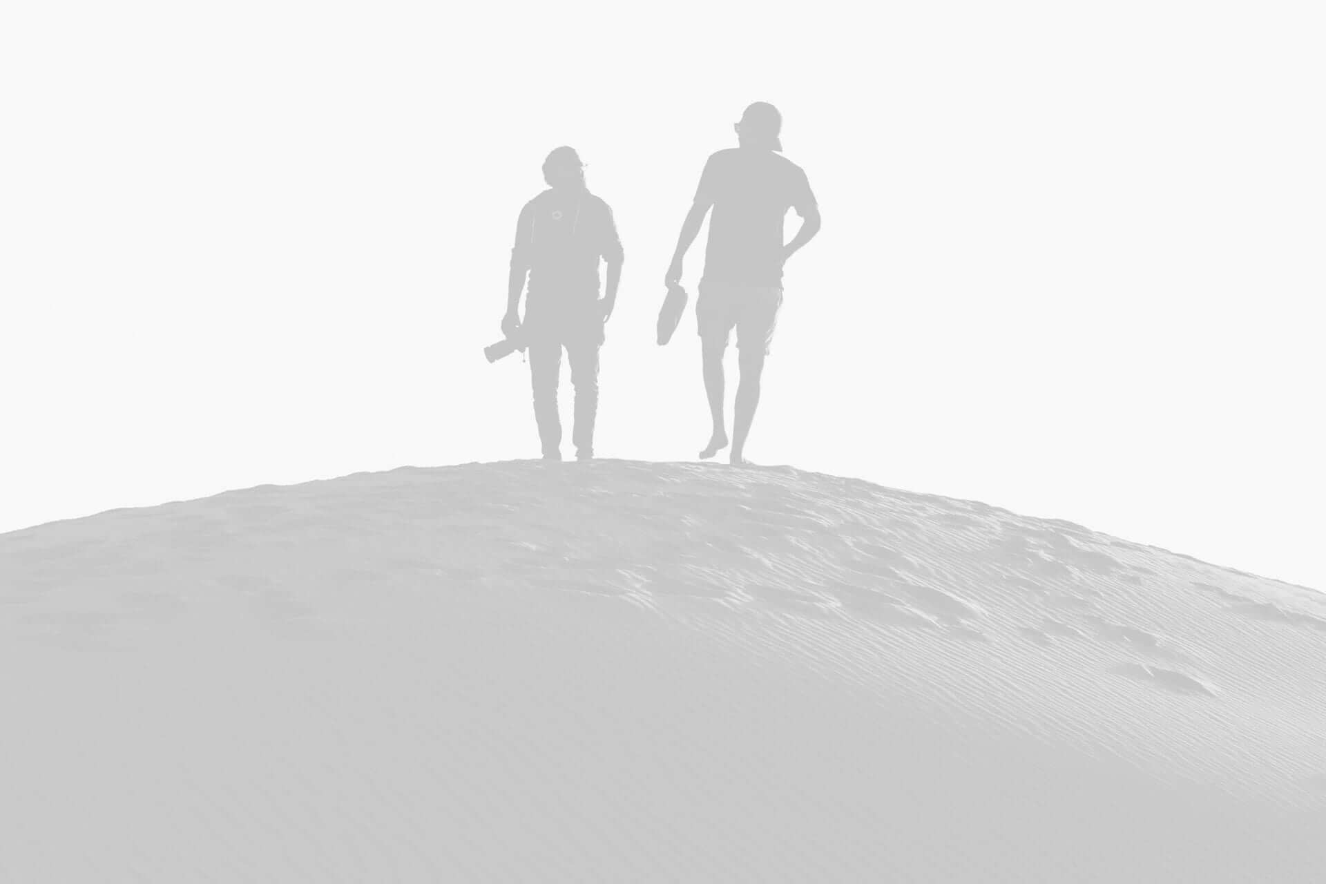 Two people standing on a sand dune, one holding a book, both silhouetted against a bright, overexposed sky.