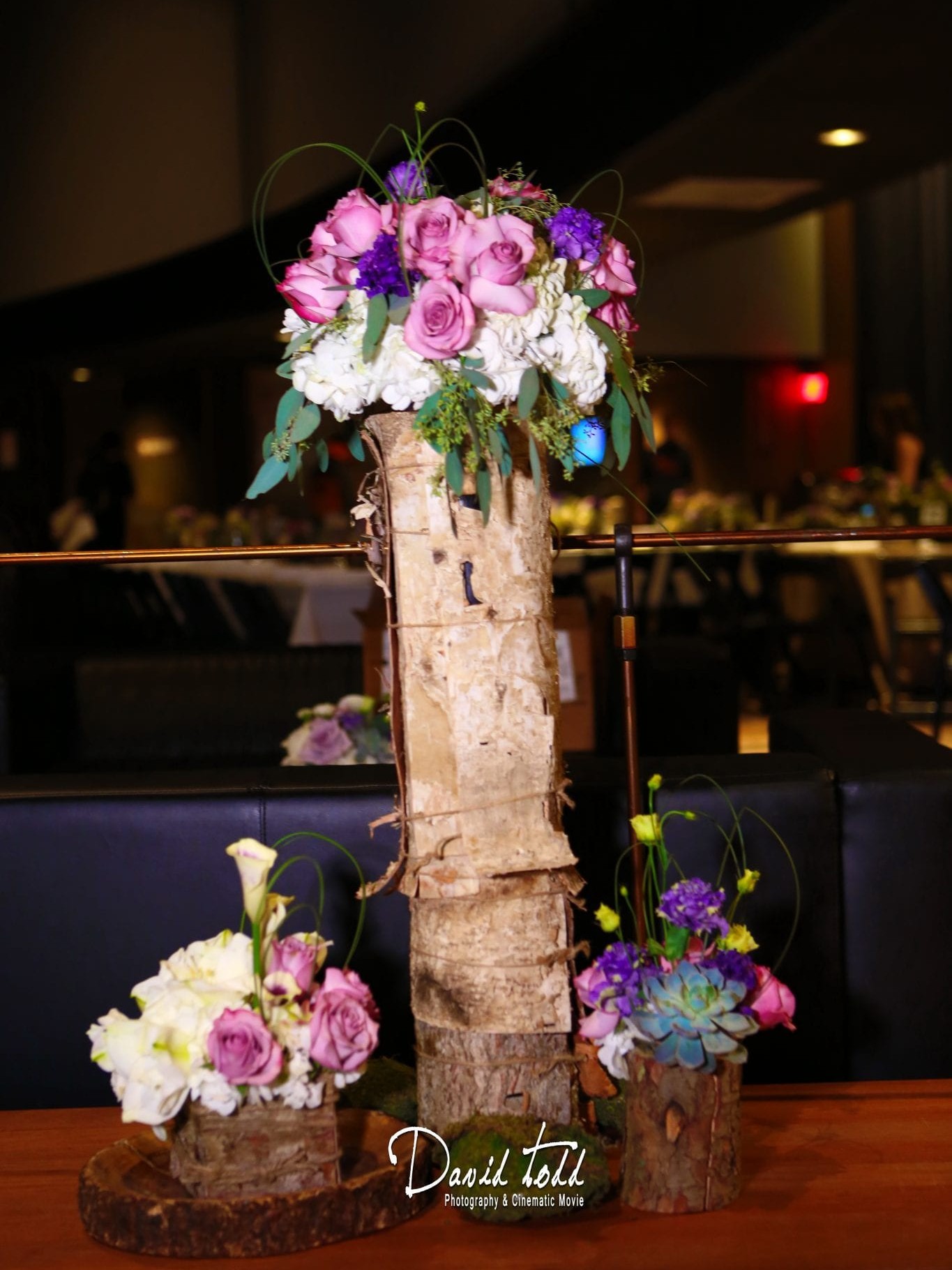A floral centerpiece on a birch log featuring pink and purple flowers, surrounded by smaller arrangements, in a dimly lit event hall.