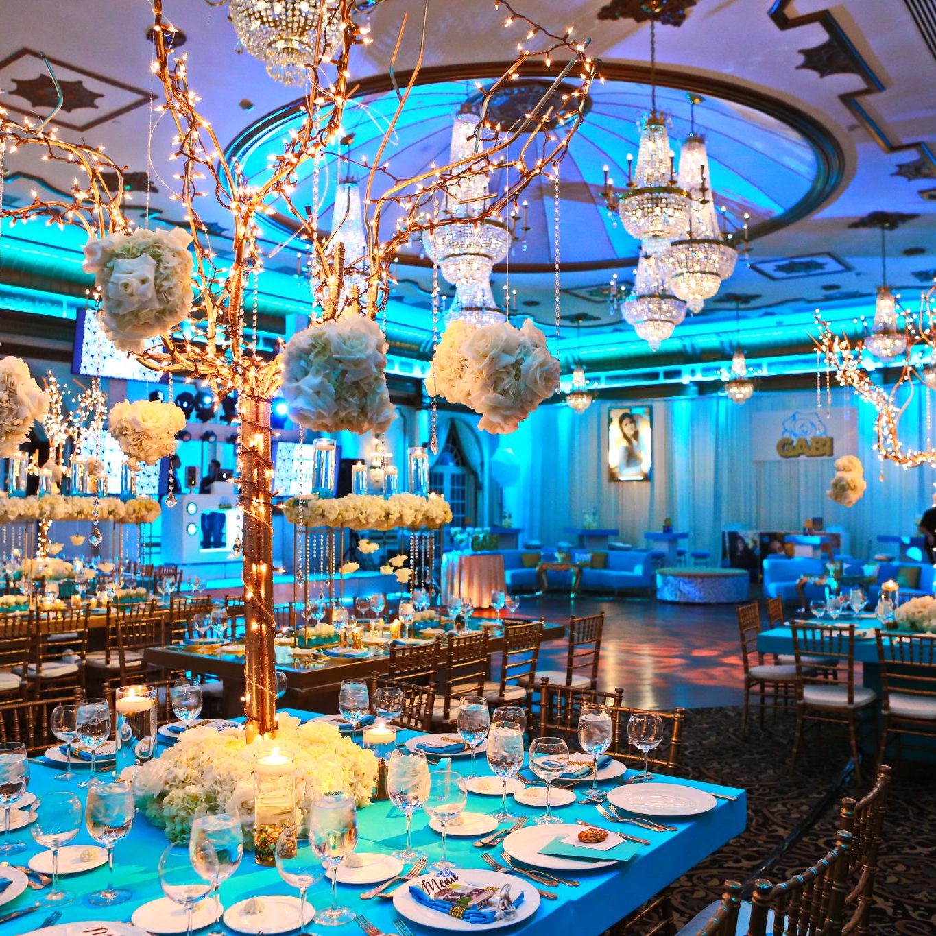 Elegant wedding reception hall decorated with blue lighting, crystal chandeliers, and floral centerpieces on dining tables.