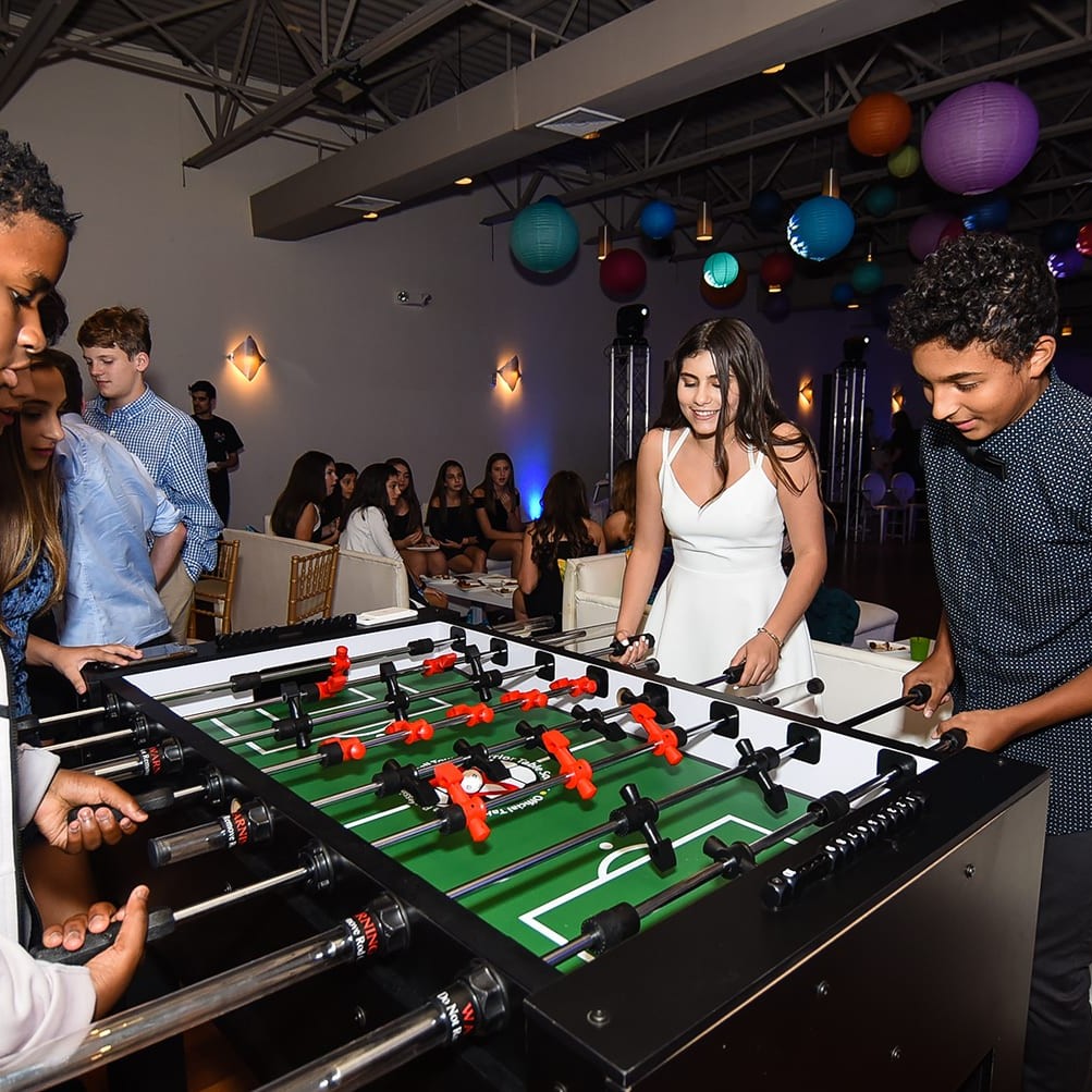 Young adults playing foosball in a lively party setting with colorful lanterns hanging above.