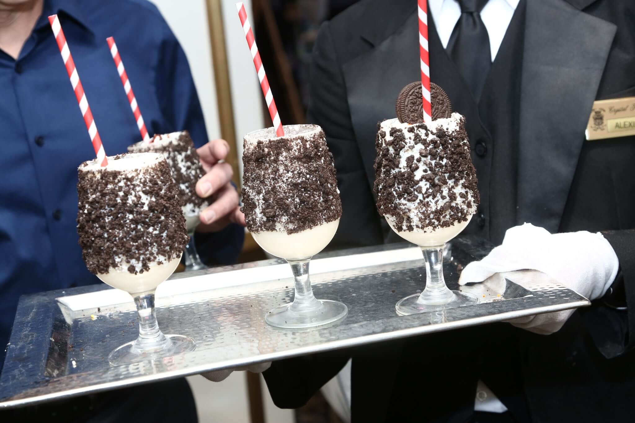 Waiter holding a tray with three extravagant chocolate milkshakes garnished with cookies and topped with whipped cream.