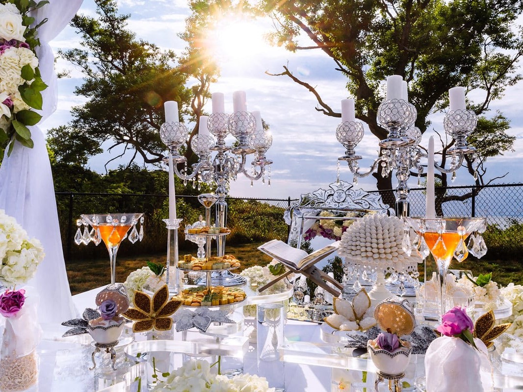 Elegant outdoor wedding table setting featuring crystal chandeliers, white flowers, and an ocean view in the background, illuminated by sunlight.