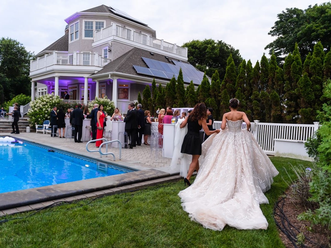 Two women in elegant dresses stand by a pool at an outdoor evening wedding reception, with guests mingling near a house.