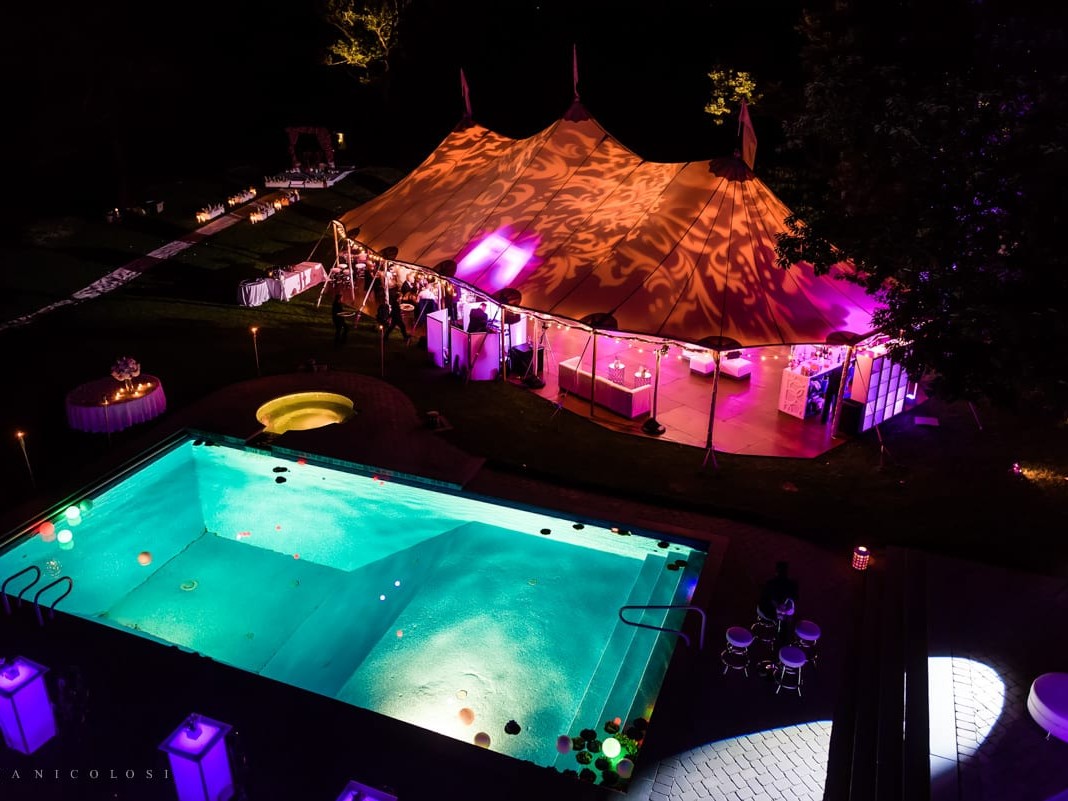 Aerial view of an outdoor evening event featuring a lit swimming pool and a large tent with colored lighting.