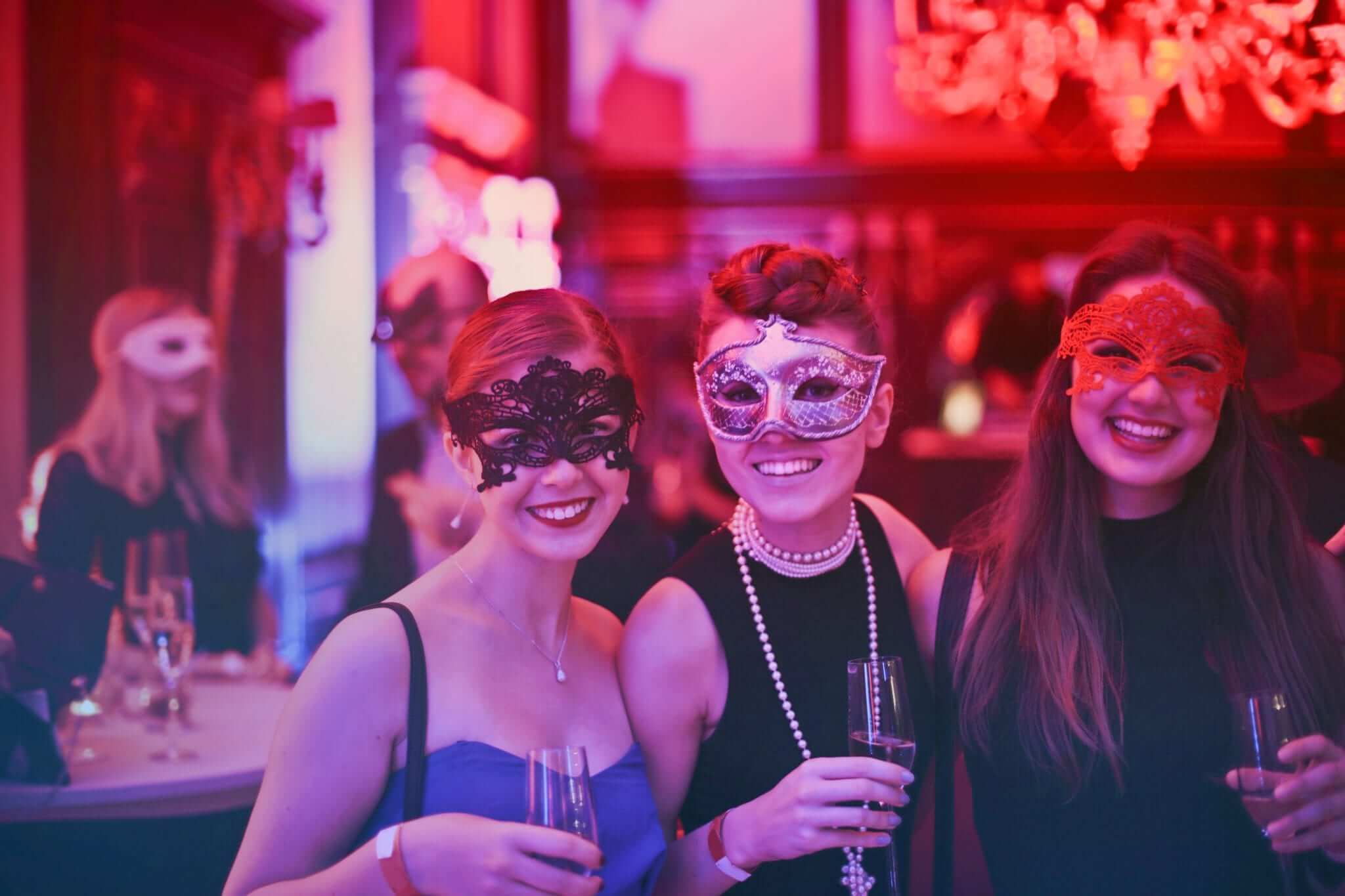Three women wearing masquerade masks and holding champagne glasses at a festive party with colorful lighting.