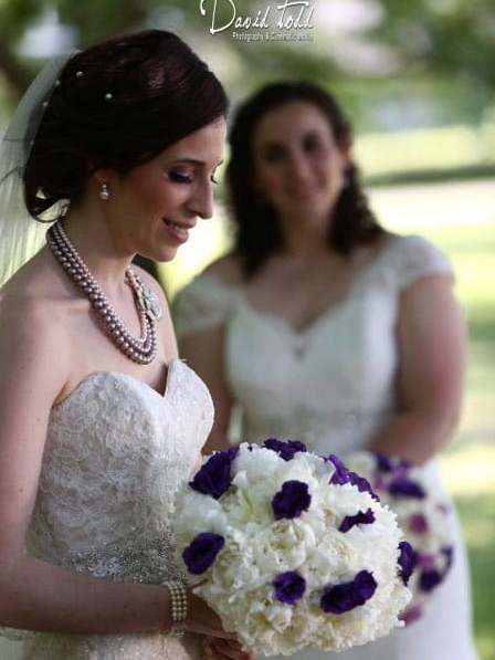 A bride holding a bouquet with purple flowers is in focus, while another woman stands blurred in the background.