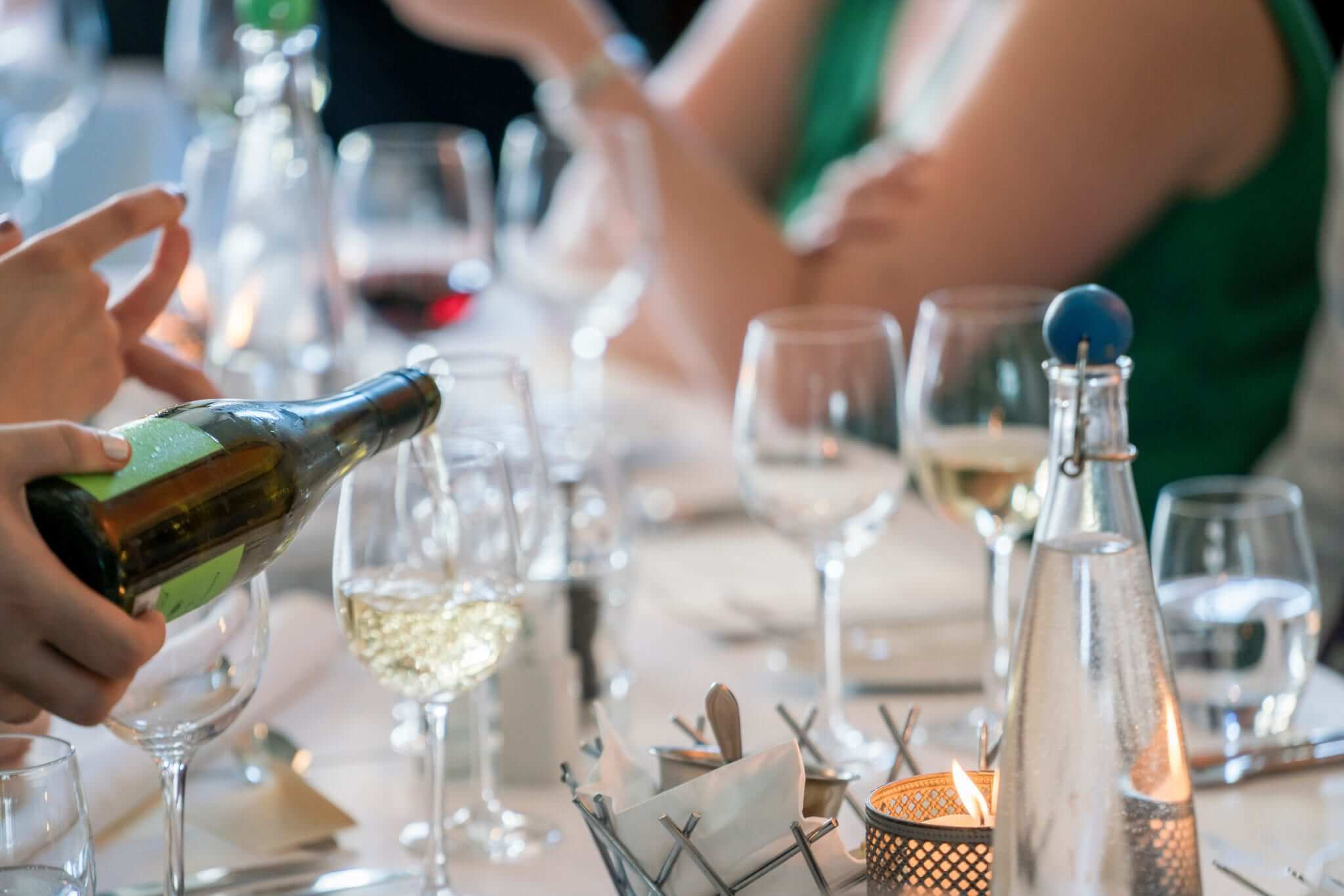 A person pours white wine into a glass at a dining table surrounded by guests.