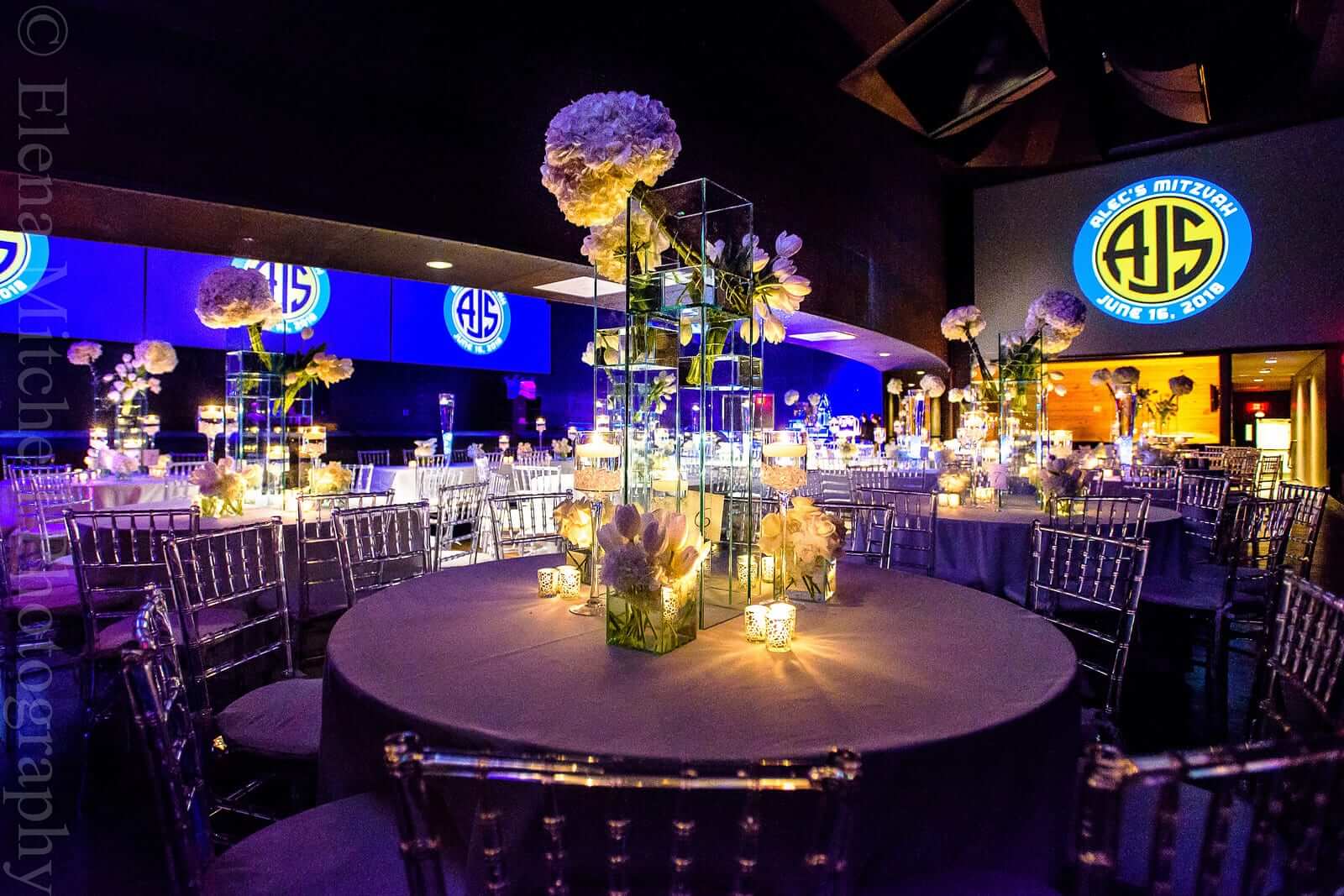 Elegant event space with round tables adorned with floral centerpieces and candles, lit by blue ambient lighting and featuring large celebratory banners with 