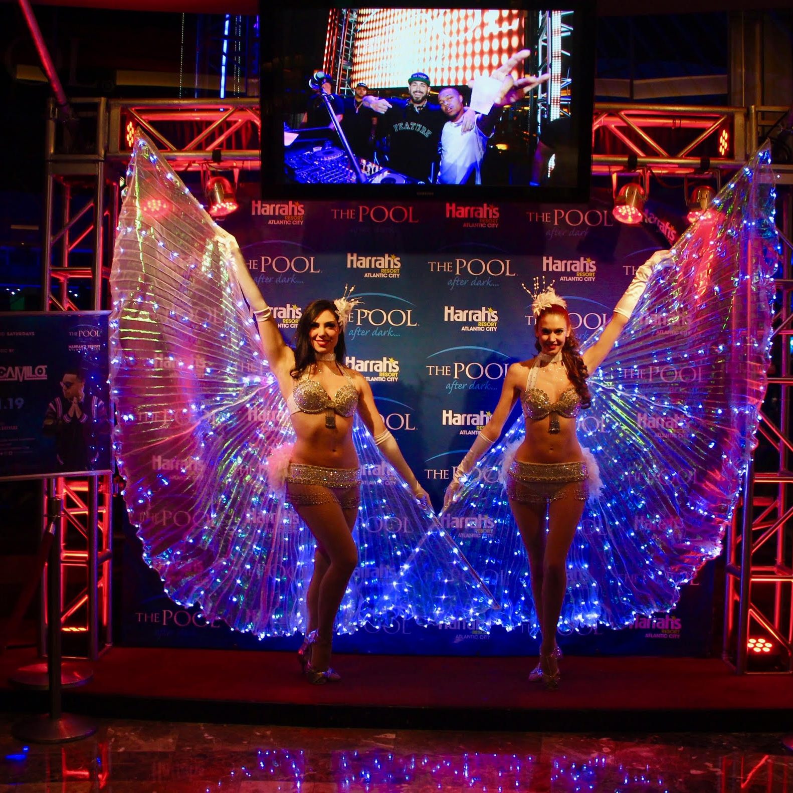 Two performers with led-lit wings stand on a stage at harrah's pool event, smiling and posing in front of a colorful backdrop.