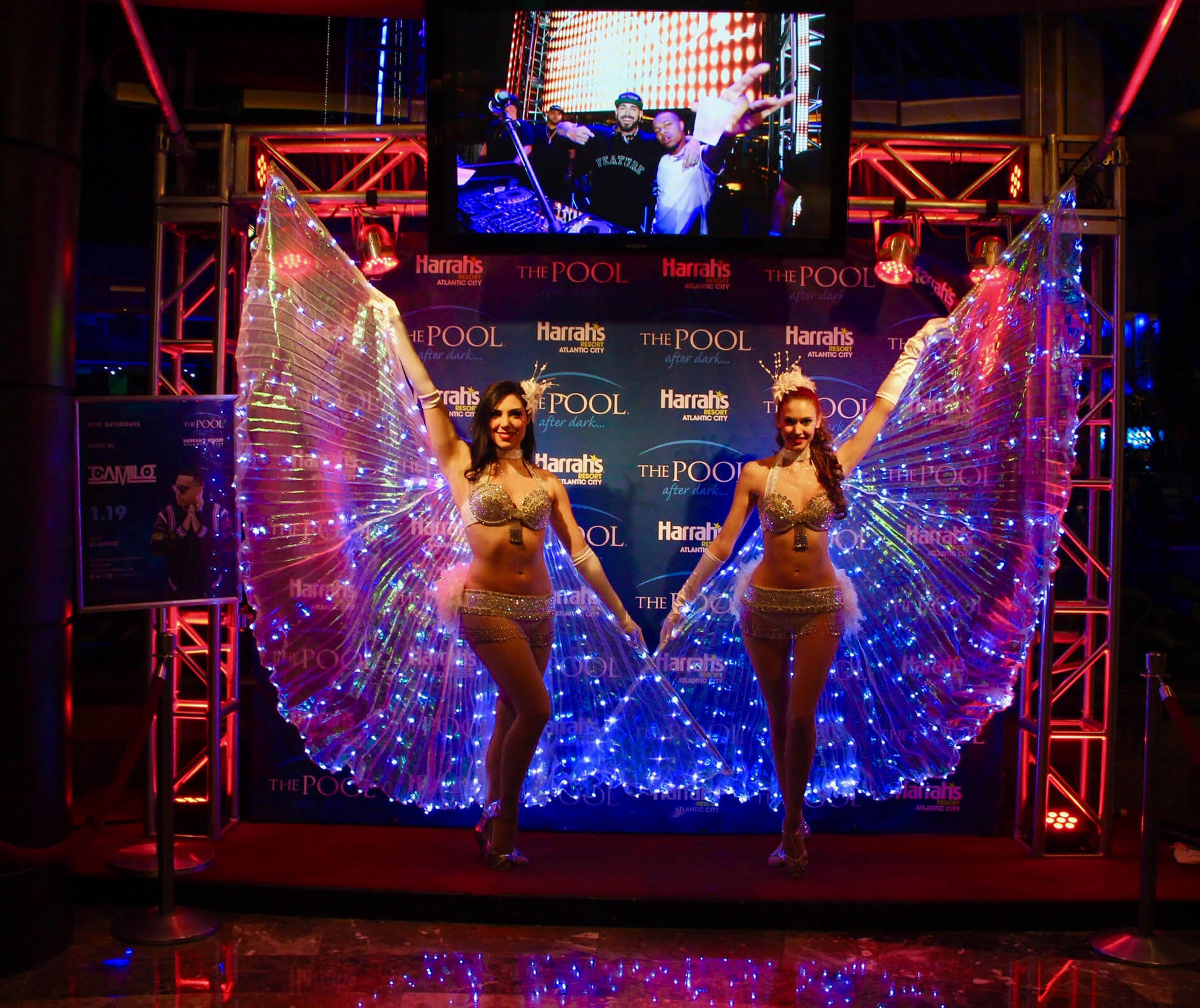 Two performers with led-lit wings stand on a stage at harrah's pool event, smiling and posing in front of a colorful backdrop.