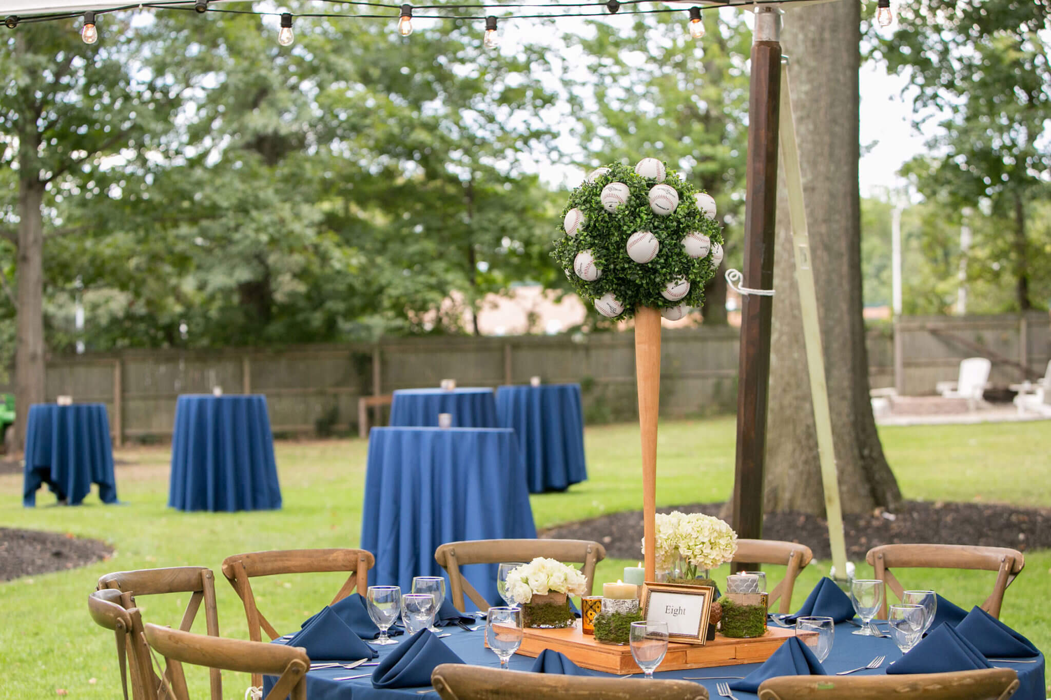 Outdoor wedding reception setup with blue tablecloths, wooden chairs, and hanging lights, decorated with white floral arrangements.