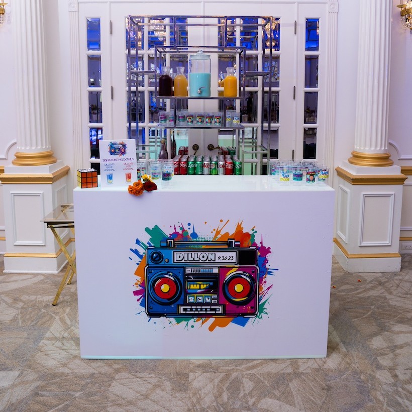 Event bar setup with a colorful boombox design panel, stocked shelves, and an arched window in the background in an elegant room.