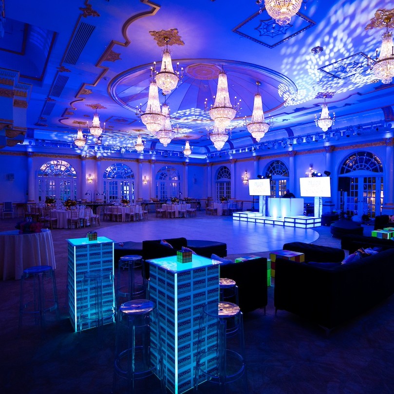Elegant event hall with blue lighting, adorned with chandeliers, set up with tables, chairs, and a central bar area.