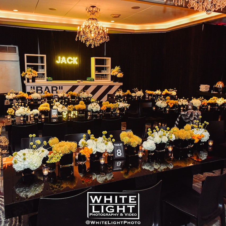 Elegant event space set up with round tables decorated with yellow and white flower centerpieces, black chairs, and a lit-up bar labeled 