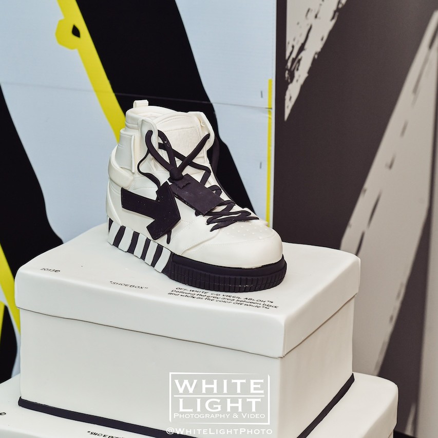 A high-top sneaker with white and black design displayed on a white pedestal against an artistic, colorfully abstract backdrop.