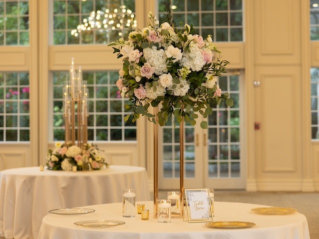 Elegant wedding reception setup with floral centerpiece on a round table, gold chargers, and candles, in a room with bright windows.