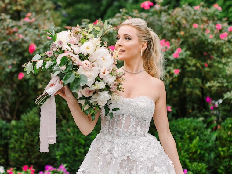 A bride in a strapless floral wedding gown holding a large bouquet, standing in a garden with pink roses in the background.