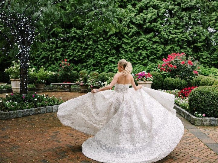A bride twirls in her voluminous wedding dress in a garden, surrounded by lush greenery and hanging lights.