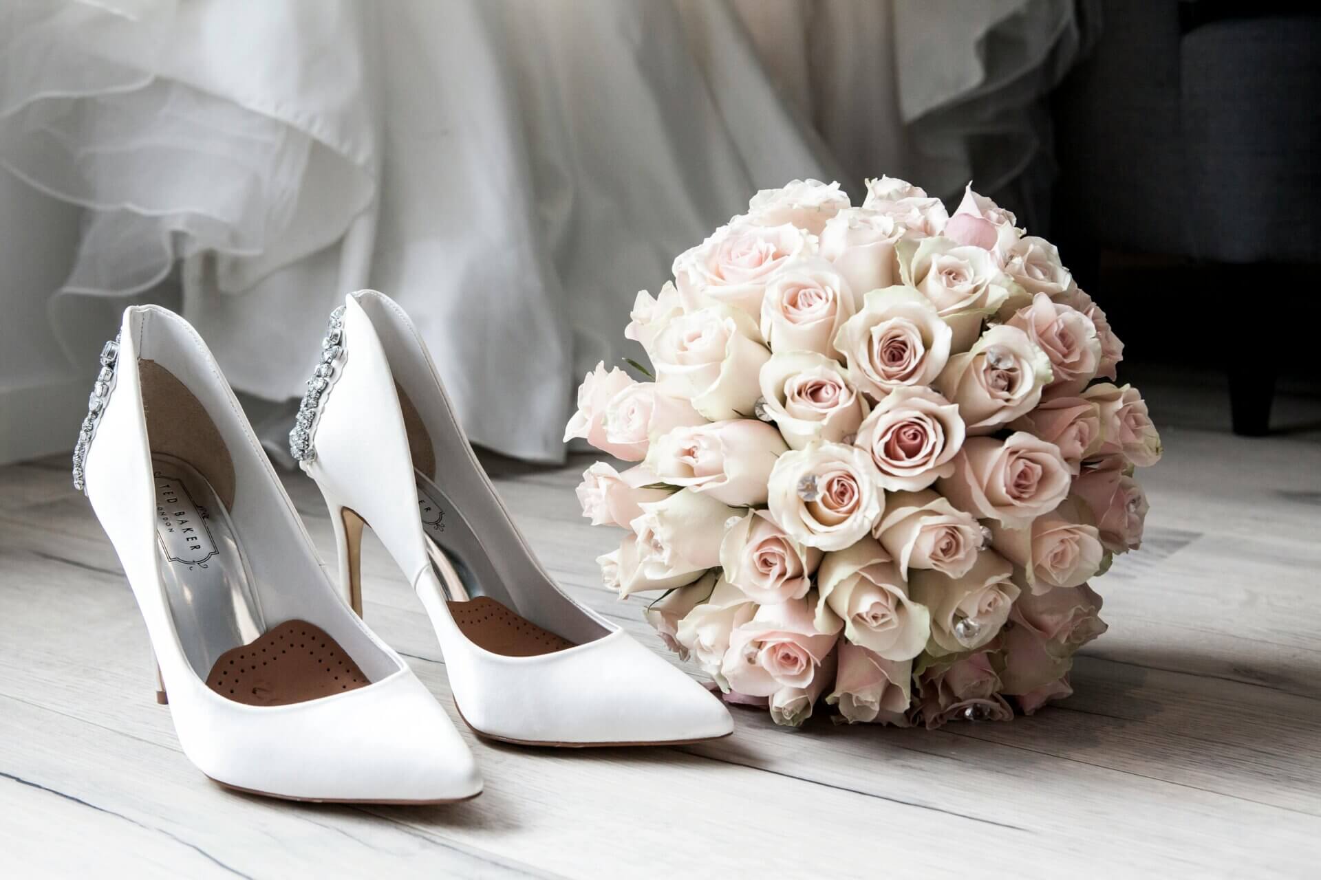 White high-heeled shoes with jeweled details rest beside a round bouquet of pale pink and white roses—perfect inspiration for your NJ wedding planning timeline—on a light wooden floor.